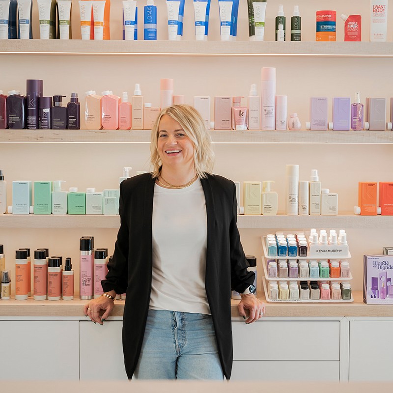 A wall full of Kevin Murphy branded hair products on wooden shelves. Owner Angie Gignac stands in front of the rack